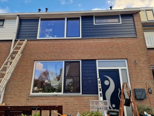 Gevel van een huis met blauwe en witte panelen, ladder en een kunstzinnig deurelement.